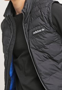 Svart pufferväst med dragkedja och en Adidas-logotyp på bröstet, med en blå invändig foder och ett quiltat mönster.