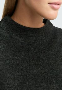 Sötét szürke kötött pulóver magas, lekerekített nyakkivágással. Közeli felvétel, amely a puha textúrát és a fonal finom eltéréseit mutatja. Minimalista kialakítás.