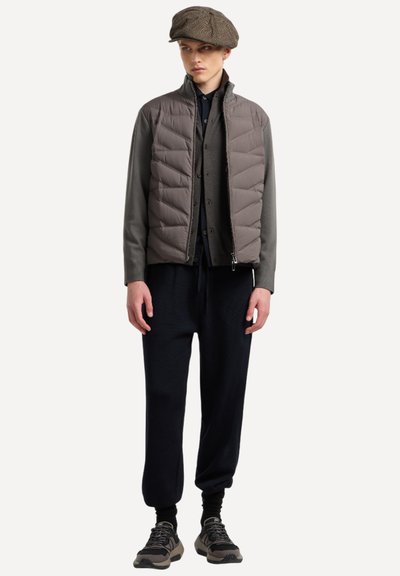 Gilet imbottito marrone tenue, con zip frontale e trapuntatura orizzontale. Abbinato a pantaloni scuri, larghi, e sneakers grigie. Accessorio: cappellino marrone.