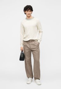 Hele beež värvi kampsun, millel on roheline ääristatud kaelus, kombineeritud laia, pruunist tekstuuriga püksipaariga. Modell hoiab väikesest musta käekotti. Valged tossud.