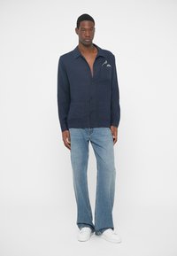 Marineblauw linnen overhemd met een relaxte pasvorm, knoopsluiting aan de voorkant en twee borstzakken. Gecombineerd met lichtblauwe wijde jeans en witte sneakers.