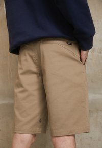 Shorts kaki à coupe droite, en tissu robuste, avec une poche latérale visible et une petite étiquette logo à l'arrière droit.