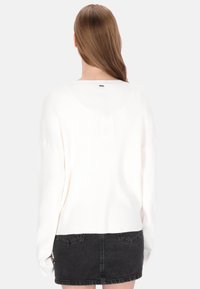 Fehér kötött pulóver laza szabással, hosszú ujjakkal és lekerekített nyakkivágással. Texturált anyag és kontrasztos fekete alsó szegély.