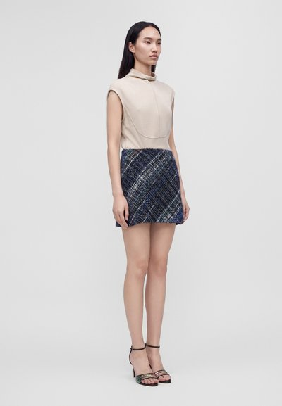 Femme aux longs cheveux foncés portant un haut beige sans manches, une mini-jupe à carreaux bleue et des sandales noires à talons hauts, debout devant un fond uni.