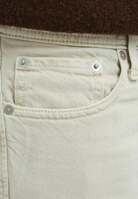 Beige denim fabric close-up with a small front pocket, silver hardware, and visible stitching details. Background is a dark brown knit texture.