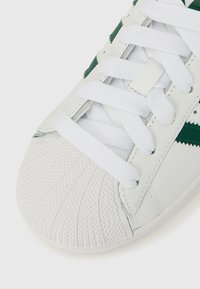 Balti ādas sporta apavi ar teksturētu gumijas pirksta daļu, baltām auklām un zaļiem akcentiem sānu panelī.