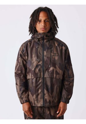 Jeune homme aux boucles aux épaules portant une veste et un pantalon camouflage marron et noir, debout devant un fond uni.