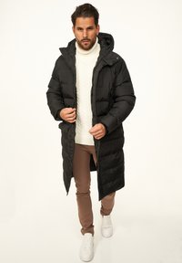 Schwarzer langer Steppmantel mit Kapuze, Reißverschluss und Seitentaschen, getragen über einem weißen, grob gestrickten Pullover und beigen Hosen.