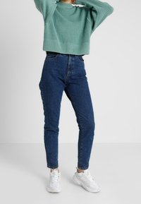 Hellgrüner Strickpullover mit geripptem Saum, kombiniert mit hochgeschnittenen dunkelblauen Jeans. Das Model trägt weiße Sneaker und steht vor einem neutralen Hintergrund.