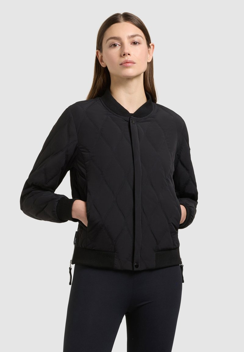 Jeune femme portant une veste bomber matelassée noire, les mains dans les poches, debout devant un fond uni gris clair.