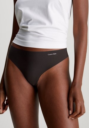 Eine Frau trägt schwarze Calvin Klein Unterwäsche und ein weißes ärmelloses Oberteil, sie steht vor einem schlichten, hellen Hintergrund.