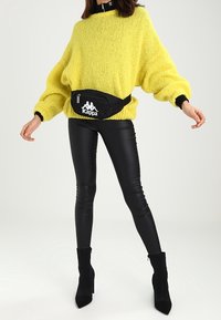 Pull jaune tricoté oversize avec accents noirs, associé à des leggings en fausse cuir noir et des bottines noires. Sac banane Kappa porté à la taille.