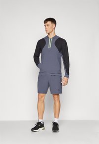 Grå atletisk hættejakke med sorte ærmer og grøn lynlåsdetalje. Kombineret med grå shorts og sorte sneakers. Enkel design med funktionelle elementer.