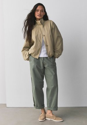 Kvinde iført beige jakke med høj krave, hvid skjorte, løse grønne bukser og beige slip-on sko, stående med hænderne i lommerne.