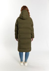 Caban lung de tip puffer, de culoare verde măsliniu, cu glugă, având cusături orizontale, croială dreaptă și mansete elastice. Purtați peste blugi albaștri și adidași.