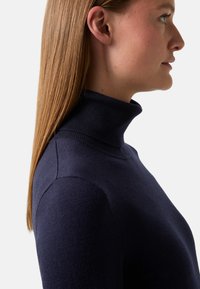 Frau mit glattem, hellbraunem Haar trägt einen navyblauen Rollkragenpullover, im Profil vor einem schlichten weißen Hintergrund dargestellt.