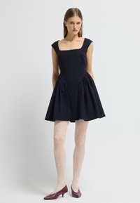 Femme debout dans une robe sans manches bleu marine à encolure carrée, avec des collants blancs à motifs et des chaussures bordeaux à talons bas, devant un fond uni.