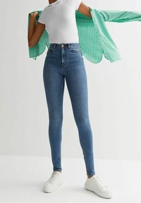 High-waisted blue jeans with a slim fit, made of denim; paired with a white T-shirt and a buttoned green and white striped shirt.