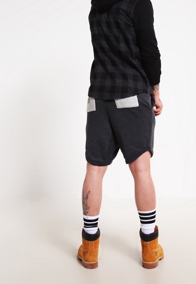 Personne portant une chemise à carreaux noire et grise, un short gris foncé avec des poches gris clair, des chaussettes rayées et des bottes de travail beige debout sur un sol blanc.