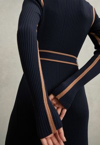 Marineblauwe geribbelde jurk met lange mouwen, voorzien van bruine strepen langs de armen en een bijpassende riem in de taille, die textuur en pasvorm benadrukt.