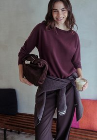 Usmívající se žena v purpurovém svetru a kalhotách drží sklenici kávy a tmavohnědou tašku uvnitř u lavičky s barevnými polštáři.