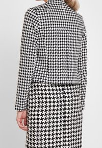 Conjunto de chaqueta y falda ajustados en patrón de ajedrez blanco y negro. Presenta mangas largas y un diseño corto. Textura de tela suave.