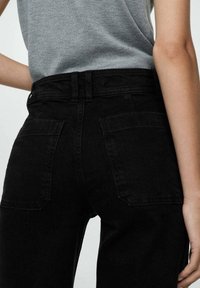 Persona que lleva unos pantalones vaqueros negros de cintura alta con bolsillos traseros y una camiseta gris de manga corta metida por dentro, mostrada de la cintura a la mitad del muslo.