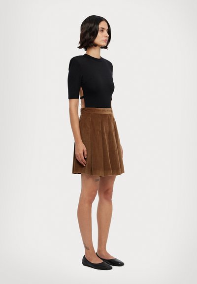 Schwarzer kurzärmeliger Bodysuit mit seitlichen Ausschnitten, kombiniert mit einem braunen Cord-Rock mit Falten. Das Modell trägt schwarze Ballerinas. Sanfte Textur.
