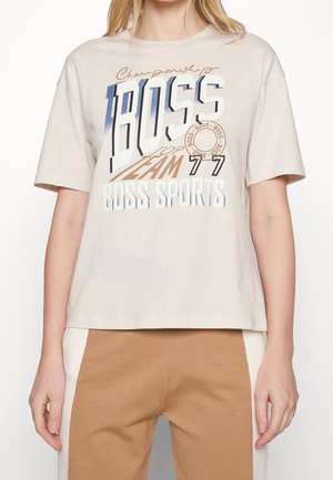 T-shirt en coton beige avec col rond, présentant un texte graphique audacieux en bleu, marron et blanc. Manches courtes, coupe décontractée et texture minimaliste.
