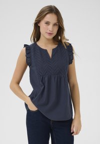 Blouse en coton bleu marine avec des manches courtes à volants et un décolleté en V profond. Présente une broderie détaillée avec un motif en chevron à l'avant.
