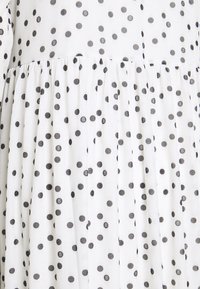Tissu blanc avec une couture froncée, décoré de pois noirs espacés régulièrement, certains contenant la lettre "B" à l'intérieur.