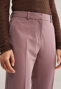 Dopasowane różowe spodnie o gładkiej teksturze, z bocznymi kieszeniami i wyraźną plisą z przodu. Noszone z brązową, teksturowaną bluzką z długim rękawem.