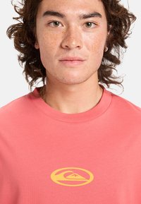 Korallfärgad t-shirt gjord av mjukt bomull; har en stor gul logotyp på bröstet med en vågdesign. Rund halsringning och korta ärmar.