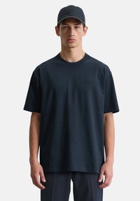 Marineblaues T-Shirt mit kurzen Ärmeln, Rundhalsausschnitt und dezentem Logodetail. Wird mit einer passenden Mütze getragen und zeigt eine lockere Passform sowie eine glatte Textur.