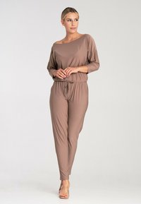 Brown jumpsuit with long sleeves, off-shoulder neckline, and elastic waistband. Smooth fabric with a relaxed fit and tapered legs. Transparent sandals.