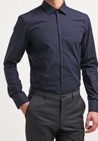 Camicia da uomo a maniche lunghe color blu navy, con design aderente, colletto button-down e texture liscia, abbinata a pantaloni grigi su misura.