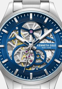 Automatyczny zegarek Kenneth Cole posiada niebieską tarczę z przezroczystym wzorem szkieletowym, srebrne wskazówki oraz stalową bransoletę.