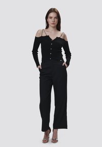 Schwarzes, geripptes Off-Shoulder-Top mit Knöpfen und Ketten-Trägern, kombiniert mit hoch taillierten schwarzen Hosen und metallischen High-Heel-Sandalen.