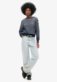 Suéter de punto azul marino con patrón de puntada abierta, puños acanalados, combinado con jeans de pierna ancha azul claro y zapatos negros gruesos.