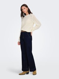 Pull en tricot crème avec motif losange texturé, associé à un jean large bleu foncé et des bottines beige. Une ceinture en cuir simple contraste avec la tenue.