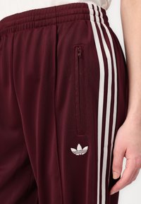 Pantaloni sportivi bordeaux cu dungi laterale albe, un buzunar cu fermoar și detalii brodate cu logo, având o textură de țesătură moale și netedă.
