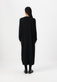 Abito lungo nero in maglia con maniche lunghe, scollo rotondo e vestibilità dritta, che scende sotto il ginocchio. Indossato con scarpe nere e calze.