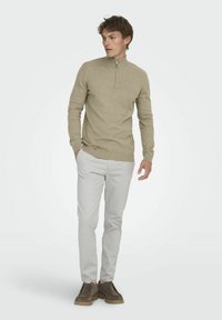 Beiger Pullover mit Reißverschluss am Hals, langen Ärmeln sowie gerippten Bündchen und Saum. Kombiniert mit hellgrauen Hosen und dunkelbraunen Schnürschuhen.