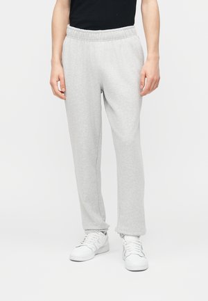 Personne portant un pantalon de survêtement gris clair, un haut noir et des baskets blanches, debout sur un fond blanc uni.