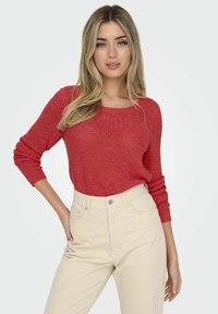 Maglione rosso lavorato a maglia con scollatura ampia e maniche lunghe, abbinato a jeans beige high-waisted. Il tessuto presenta un motivo testurizzato.