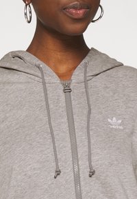 Sweat à capuche gris à fermeture éclair en tissu doux avec une capuche, équipé de cordons de serrage et d'un petit logo adidas blanc sur la poitrine gauche.