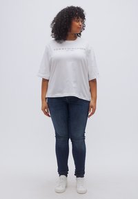 Λευκό κοντομάνικο T-shirt με το λογότυπο "TOMMY HILFIGER", σε συνδυασμό με σκούρο μπλε τζιν και λευκά αθλητικά παπούτσια. Μπούκλες μαλλιών, σε όρθια στάση.