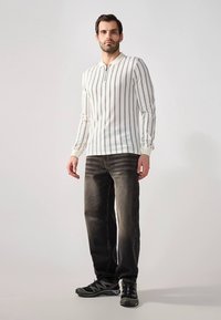 Hosszú ujjú fehér ing függőleges fekete csíkokkal, gombos nyakkivágással, sötét farmerrel és fekete sportcipővel párosítva. Egyszerű kialakítás, testhez simuló szabás.