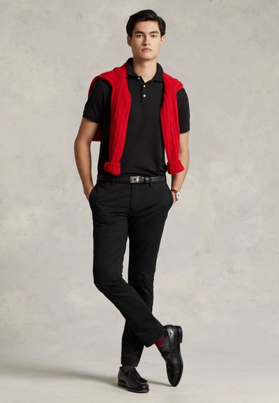 Mann steht mit Händen in den Taschen, trägt ein schwarzes Polohemd, schwarze Hose, schwarze Schuhe, einen roten Pullover über den Schultern und eine Uhr am linken Handgelenk.
