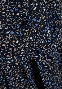 Tissu bleu marine avec un motif floral en crème, rose et bleu ; présente un détail de bouton et une texture plissée près de l'ourlet.